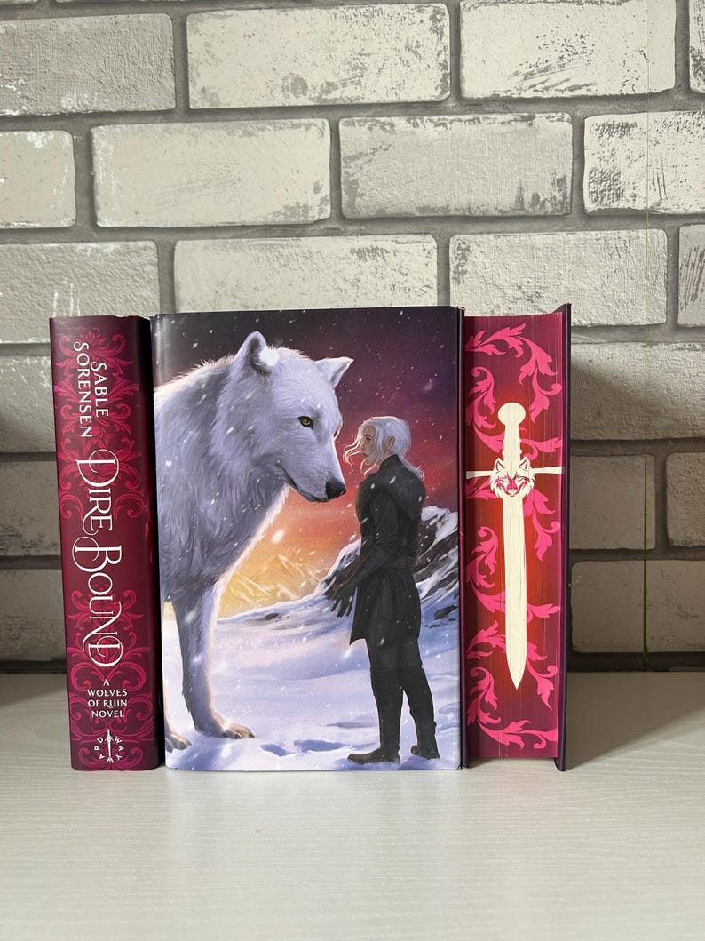 May include: A book cover featuring a white wolf facing a person in a snowy landscape. The book is titled "Dire Bound" by Sable Sorensen. The spine is burgundy with white text. The cover art depicts a fantasy theme.