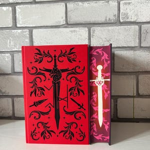 May include: A red book with black decorative elements and a sword design. The spine of the book is visible, featuring a white sword design against a pink and purple background. The book is set against a white surface and a brick wall.