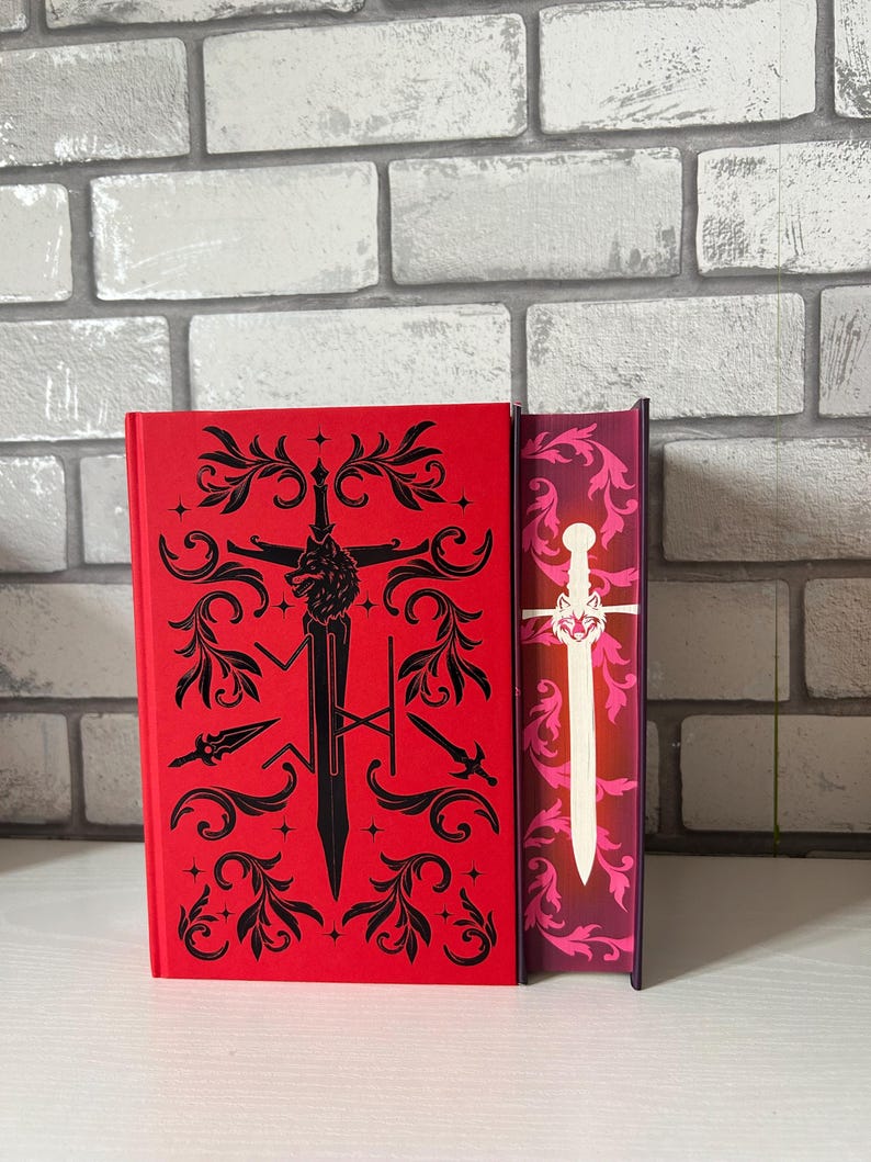 May include: A red book with black decorative elements and a sword design. The spine of the book is visible, featuring a white sword design against a pink and purple background. The book is set against a white surface and a brick wall.