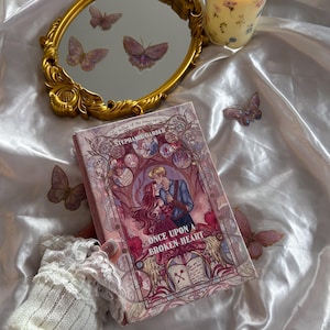 Può includere: Uno specchio con cornice dorata con adesivi a farfalla, una candela e un libro intitolato "Once Upon a Broken Heart" di Stephanie Garber. La copertina del libro presenta una coppia in una scena romantica. La scena è ambientata su una superficie di raso bianco.