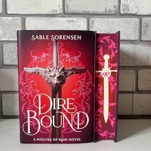 May include: A hardcover book titled "Dire Bound" by Sable Sorensen. The cover is predominantly red with a silver sword design and a wolf head. The spine is visible, featuring a matching design with a white sword.