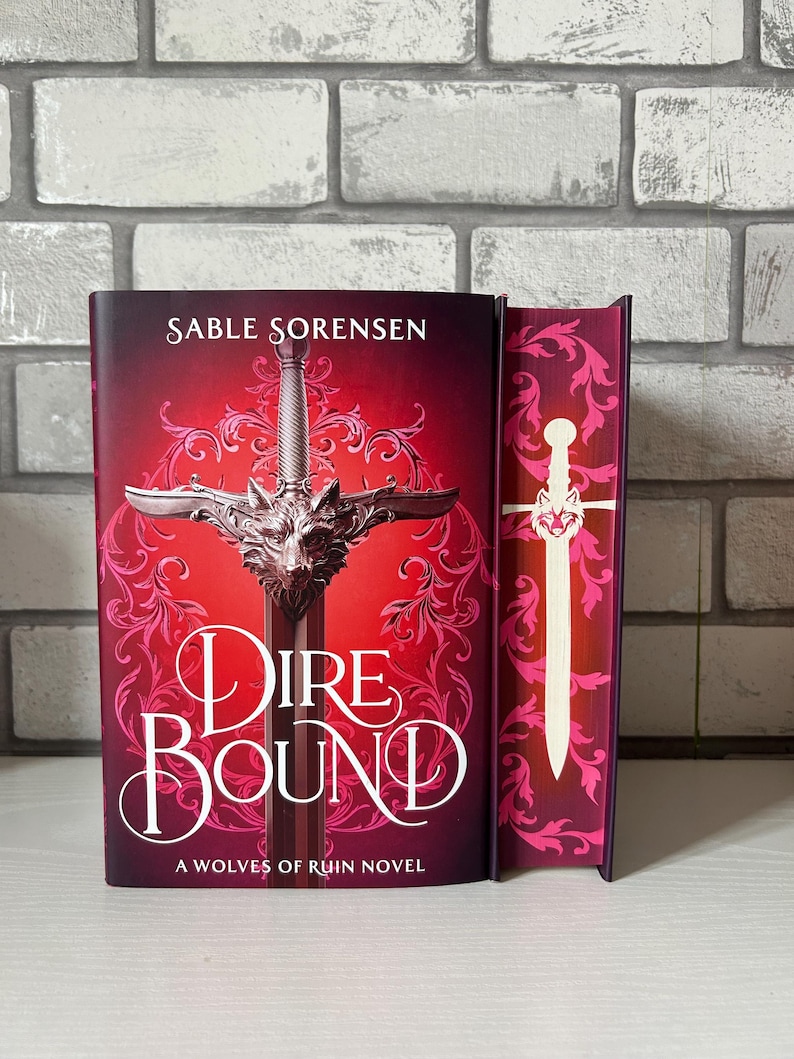 May include: A hardcover book titled "Dire Bound" by Sable Sorensen. The cover is predominantly red with a silver sword design and a wolf head. The spine is visible, featuring a matching design with a white sword.