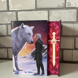 May include: Two hardcover books. The first book features a white wolf and a person in black clothing in a snowy landscape. The second book has a red cover with a white sword and floral design. The books are displayed upright.