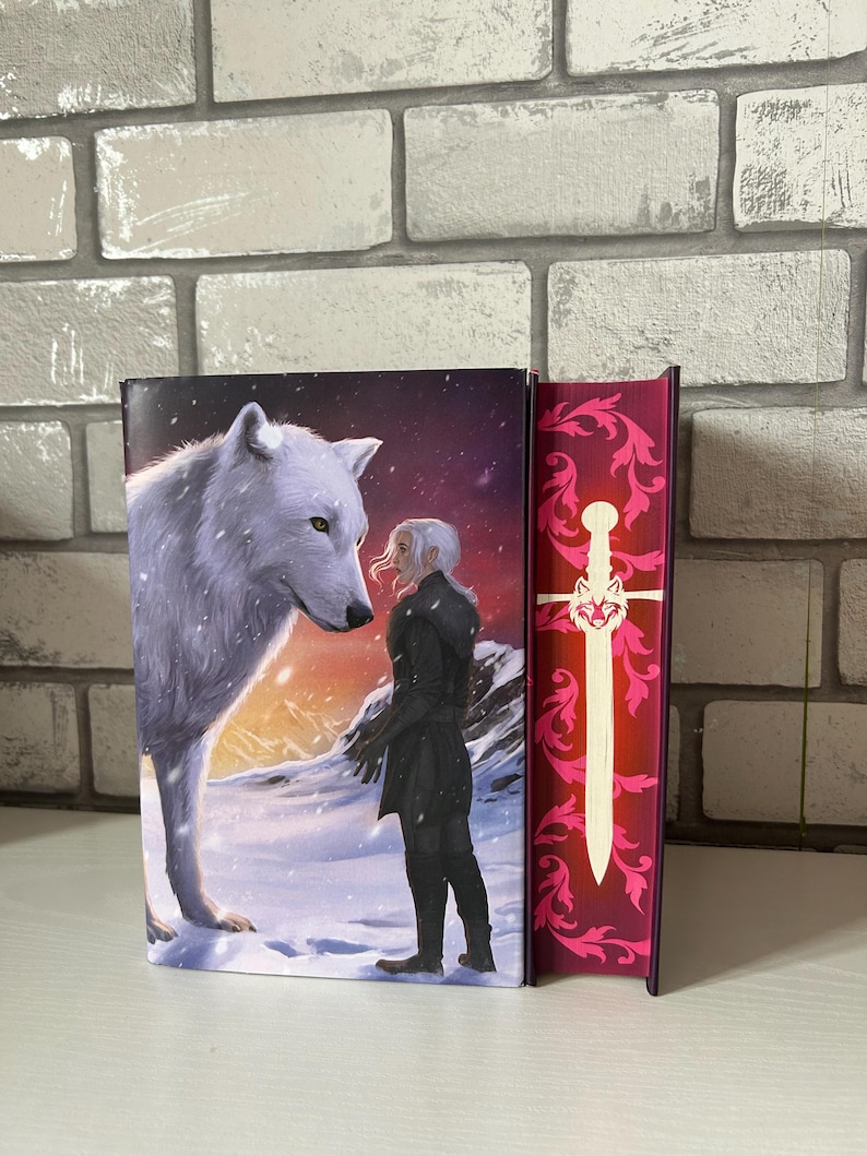 May include: Two hardcover books. The first book features a white wolf and a person in black clothing in a snowy landscape. The second book has a red cover with a white sword and floral design. The books are displayed upright.