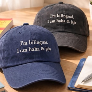 May include: Two baseball caps, one navy blue and one charcoal gray, are displayed on a wooden table. Both caps have the text "I'm bilingual, I can haha & jaja" embroidered in white. Other items on the table include a mug, books, and a pen.