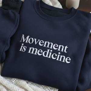 May include: Navy blue sweatshirt with the text "Movement is medicine" in white. The sweatshirt is folded on a cream-colored knitted blanket. A small vase with greenery and an open book are in the background.