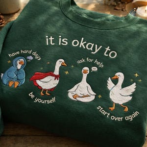 May include: Dark green sweatshirt with embroidered geese illustrations. The text "it is okay to" is above the geese, with phrases like "have hard days," "be yourself," "ask for help," and "start over again" below the geese. The geese are in various poses.