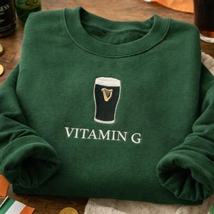 May include: A dark green sweatshirt with a crew neck and long sleeves. The sweatshirt features an embroidered design of a pint of stout with the text "VITAMIN G" in white. The image also includes a pint of stout, Guinness bottles, and Irish flags.