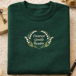 May include: A dark green sweatshirt with the embroidered text "Dearest Gentle Reader" surrounded by a wreath of white flowers and green leaves. The sweatshirt is folded neatly on a lace surface.