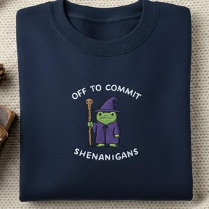 May include: Navy blue sweatshirt with the phrase "OFF TO COMMIT SHENANIGANS" and a cartoon frog dressed as a wizard. The frog is wearing a purple robe and hat, holding a staff. The sweatshirt is folded on a cream-colored surface.