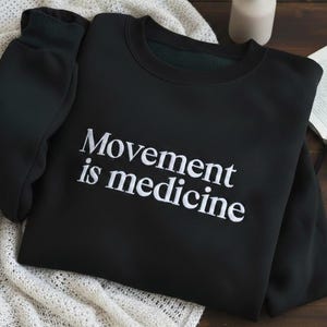 May include: Black crew neck sweatshirt with the white text "Movement is medicine". The sweatshirt is folded on a cream knitted blanket. A small white vase and a book are in the background. The sweatshirt has long sleeves.