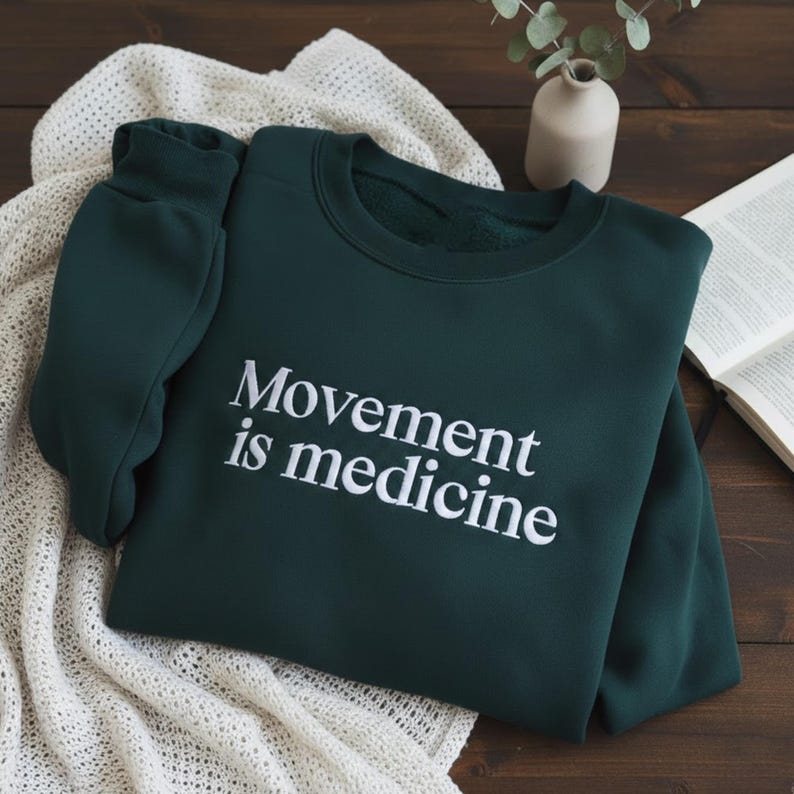 May include: A dark green sweatshirt with the slogan "Movement is medicine" in white. The sweatshirt is folded on a cream knitted blanket. A small white vase and a book are in the background.