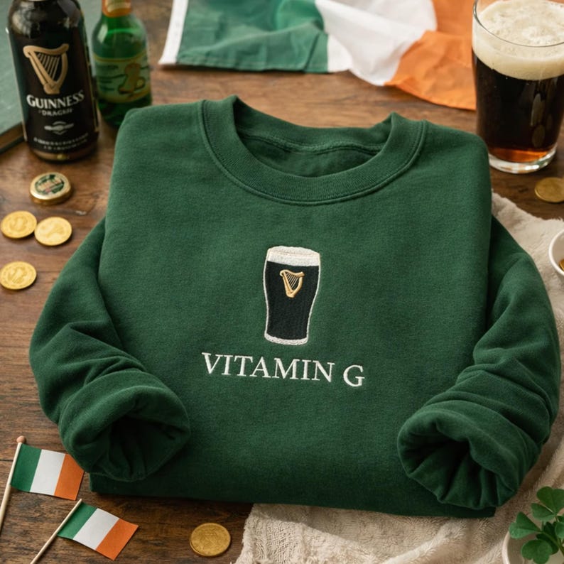May include: A dark green sweatshirt with a crew neck and long sleeves. The sweatshirt features an embroidered design of a pint of stout with the text "VITAMIN G" in white. The image also includes a pint of stout, Guinness bottles, and Irish flags.