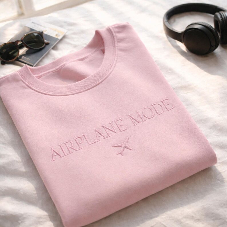 May include: A light pink sweatshirt with the words "AIRPLANE MODE" embroidered across the chest, along with a small airplane symbol. The sweatshirt is folded on a white surface, with sunglasses and headphones in the background.