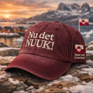 May include: A maroon baseball cap with white embroidered text "Nu det NUUK!" and a Greenland flag patch. The cap has a curved brim and is set against a scenic backdrop of mountains and water.