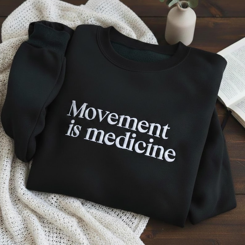 May include: Black crew neck sweatshirt with the white text "Movement is medicine". The sweatshirt is folded on a cream knitted blanket. A small white vase and a book are in the background. The sweatshirt has long sleeves.