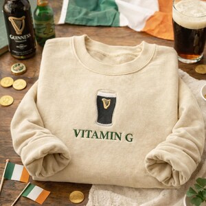 May include: A beige sweatshirt with a black and gold embroidered pint of Guinness and the words "VITAMIN G" in green. The image includes bottles of Guinness, a pint of beer, and Irish flags.