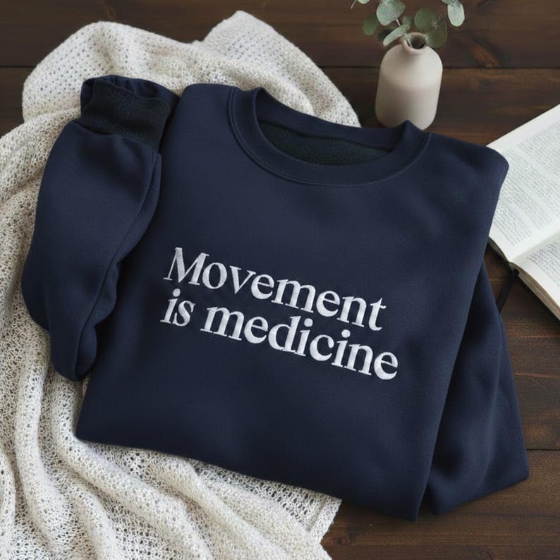 May include: Navy blue sweatshirt with the text "Movement is medicine" in white. The sweatshirt is folded on a cream-colored knitted blanket. A small vase with greenery and an open book are in the background.