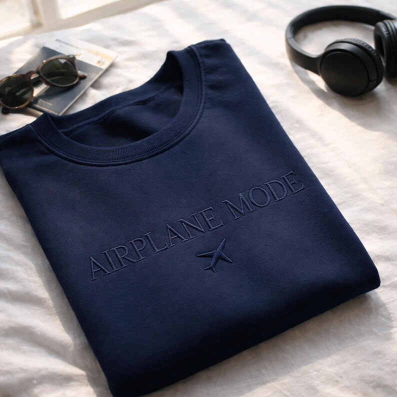 May include: Navy blue sweatshirt with the embroidered text "AIRPLANE MODE" and a small airplane icon. The crew neck sweatshirt is folded on a white surface, with sunglasses and headphones in the background.