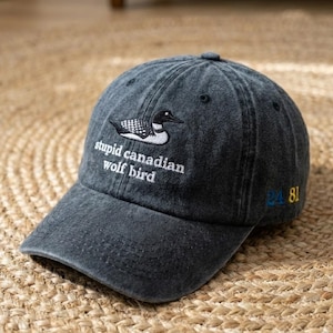 May include: A dark gray baseball cap with the text "stupid canadian wolf bird" embroidered in white. A black and white bird graphic is above the text. The number "24 81" is embroidered on the side in blue and yellow.
