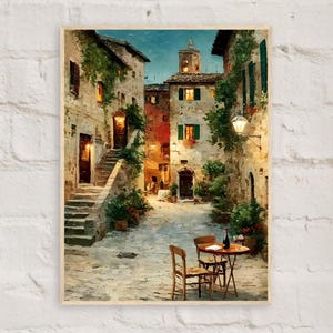 May include: Oil painting of a European village scene. The artwork depicts stone buildings with arched doorways, windows with green shutters, and climbing vines. A small table and chairs are set in the cobblestone square. The painting is framed in a light wood.