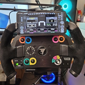 Può includere: Un volante da corsa Thrustmaster T300 nero con uno smartphone che mostra i dati di telemetria di gara. Il volante presenta pulsanti colorati e un'impugnatura simile alla pelle scamosciata. Lo schermo mostra velocità, tempi sul giro e altre metriche.