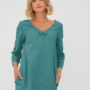 Könnte beinhalten: Türkisfarbenes Kleid mit V-Ausschnitt und kleiner Schleife. Das Kleid hat Dreiviertelärmel und zwei Vordertaschen. Der Stoff hat eine strukturierte Optik und das Kleid reicht bis zu den Knien.