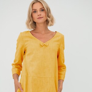 Könnte beinhalten: Ein gelbes Leinenkleid mit V-Ausschnitt und einer Schleife am Ausschnitt. Das Kleid hat 3/4-Ärmel und Taschen an den Seiten.