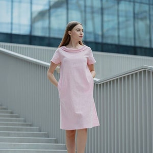 May include: A woman wearing a pink linen dress with a collared neckline and short sleeves. She is standing on a set of stairs in front of a modern building.