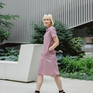 Könnte beinhalten: Eine Frau in einem rosafarbenen Leinenkleid mit kurzen Ärmeln und Taschen, die in einer modernen Umgebung mit einer weißen Bank und grünen Pflanzen steht.