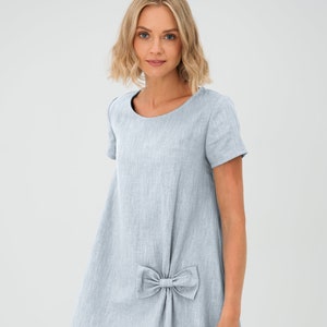 May include: A light blue linen dress with a bow detail. The dress features short sleeves and a round neckline. The knee-length dress has a loose, flowing silhouette, suitable for casual wear. The fabric appears to be lightweight and breathable.
