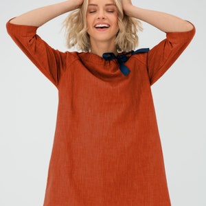 May include: A rust-colored dress with three-quarter sleeves and a loose fit. A dark blue bow adorns the neckline. The dress is made of a textured fabric and has a simple, elegant design, suitable for casual wear.