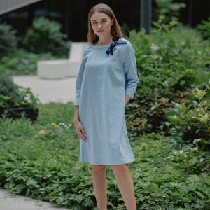 Könnte beinhalten: Eine Frau trägt ein hellblaues Leinenkleid mit einer schwarzen Schleife am Ausschnitt. Sie steht auf einem Betonweg mit grünen Pflanzen im Hintergrund. Sie trägt weiße Turnschuhe.