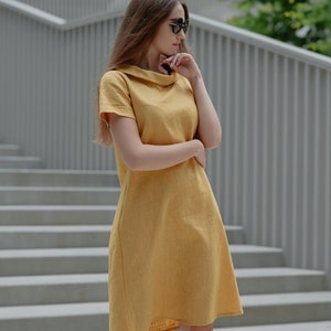May include: A woman wearing a yellow linen dress with a collared neckline and short sleeves. She is standing on a set of concrete stairs.