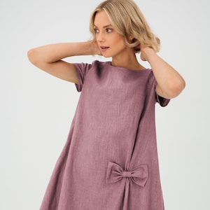 Könnte beinhalten: Ein mauvefarbenes Leinenkleid mit einer Schleife an der Seite. Das Kleid hat kurze Ärmel und eine lockere Passform.