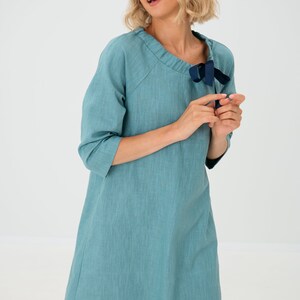 Könnte beinhalten: Türkisfarbenes Kleid mit Dreiviertelärmeln und einem einzigartigen Ausschnitt. Eine dunkelblaue Schleife schmückt den Ausschnitt. Das Kleid hat ein schlichtes, fließendes Design, geeignet für lässige oder halbformelle Anlässe.