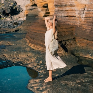 May include: A woman in a cream-colored linen jumpsuit stands on a rocky shore, holding a woven tote bag. The background features layered rock formations in shades of orange and brown under a clear blue sky. The scene evokes a sense of natural beauty and tranquility.