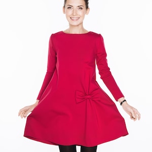 Könnte beinhalten: Ein leuchtend rotes Kleid mit langen Ärmeln und einer Schleife auf der Vorderseite. Das Kleid hat einen ausgestellten Rock und wird mit schwarzen Strumpfhosen getragen. Das Model lächelt vor weißem Hintergrund.