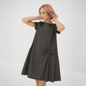 Könnte beinhalten: Ein dunkelgraues, kurzärmeliges Kleid mit einer Schleife an der Seite. Das Kleid hat eine lockere Passform und einen leicht ausgestellten Rock.