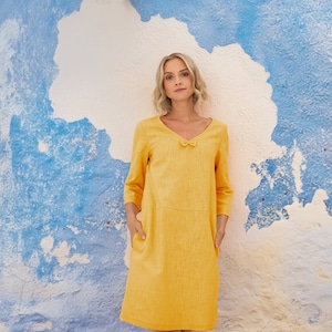 Könnte beinhalten: Ein gelbes Leinenkleid mit V-Ausschnitt und einer Schleife am Ausschnitt. Das Kleid hat Dreiviertelärmel und Taschen an den Seiten.