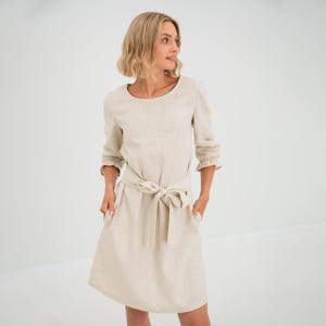 Könnte beinhalten: Ein beiges Leinenkleid mit einer Schleife in der Taille und 3/4 Ärmeln. Das Kleid hat einen runden Ausschnitt und Taschen an den Seiten.