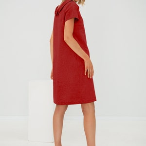 May include: A woman wearing a red linen dress with a large collar. The dress has short sleeves and a straight silhouette. She is wearing black leather flats.