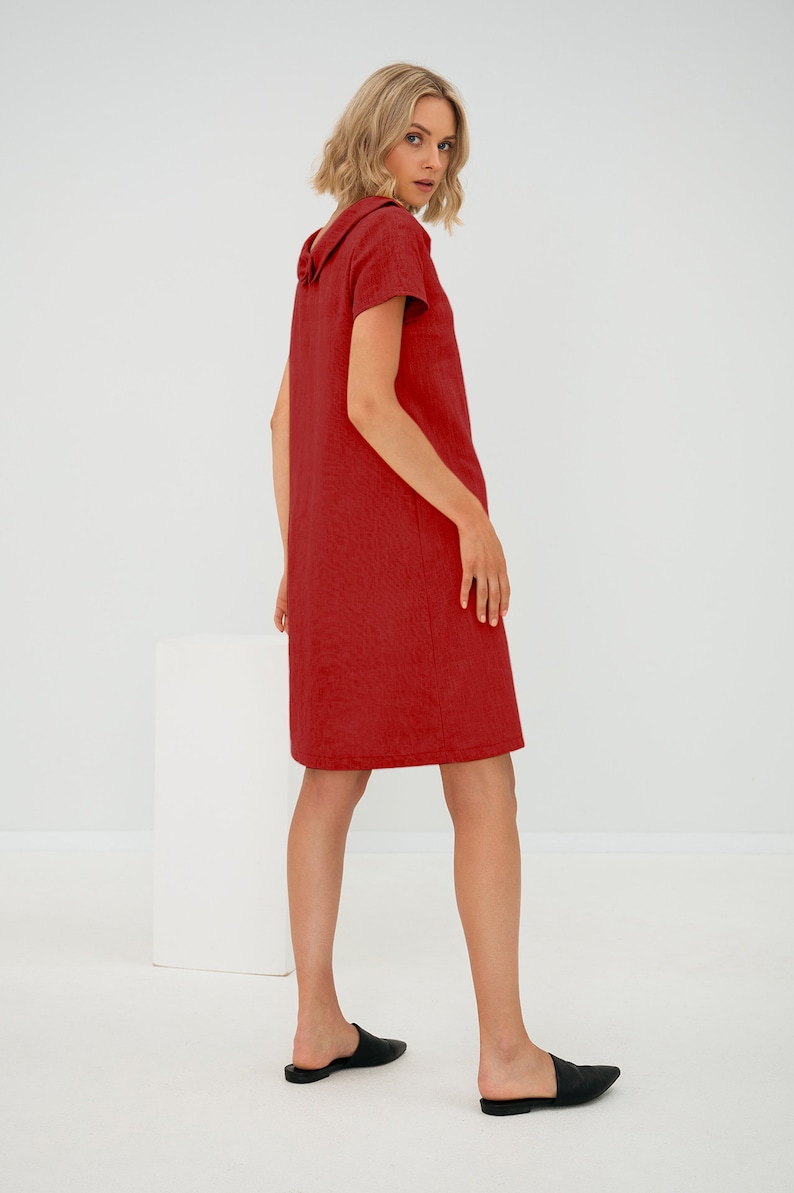 May include: A woman wearing a red linen dress with a large collar. The dress has short sleeves and a straight silhouette. She is wearing black leather flats.