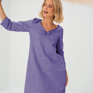 Könnte beinhalten: Ein fliederfarbenes Kleid mit V-Ausschnitt und Dreiviertelärmeln. Das Kleid hat eine kleine Schleife am Ausschnitt und eine schlichte A-Linien-Silhouette. Der Stoff hat ein strukturiertes Aussehen.