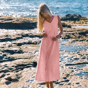May include: A sleeveless, light pink dress with a V-neck and a matching waist tie. The dress is ankle-length and made of a lightweight fabric. The background shows a rocky beach and the ocean.