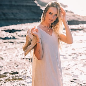 Puede incluir: Vestido beige claro con cuello en V. El vestido es sin mangas y llega hasta los tobillos. Un sombrero de paja se sostiene en el brazo. El fondo presenta una costa rocosa y acantilados.