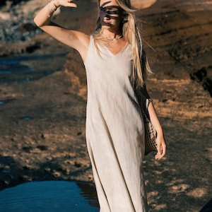 May include: A light beige, V-neck maxi dress with spaghetti straps. The dress is made of a lightweight fabric and falls to the ankles. A woven bag hangs from the shoulder. The person is standing on a rocky surface.