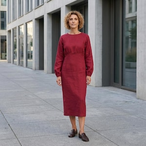 May include: A long-sleeved midi dress in a deep red color. The dress features a round neckline, a fitted bodice with a defined waistline, and a straight skirt that falls below the knees. The sleeves are long with gathered cuffs at the wrists.