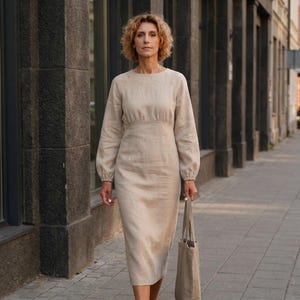 May include: A beige midi-length dress with long sleeves, a round neckline, and a fitted bodice that transitions into a straight skirt. The garment features a subtle gathered detail at the waistline and is paired with a matching beige tote bag.