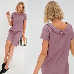 Könnte beinhalten: Ein mauvefarbenes Leinenkleid mit Peter-Pan-Kragen und einer Schleife auf dem Rücken.